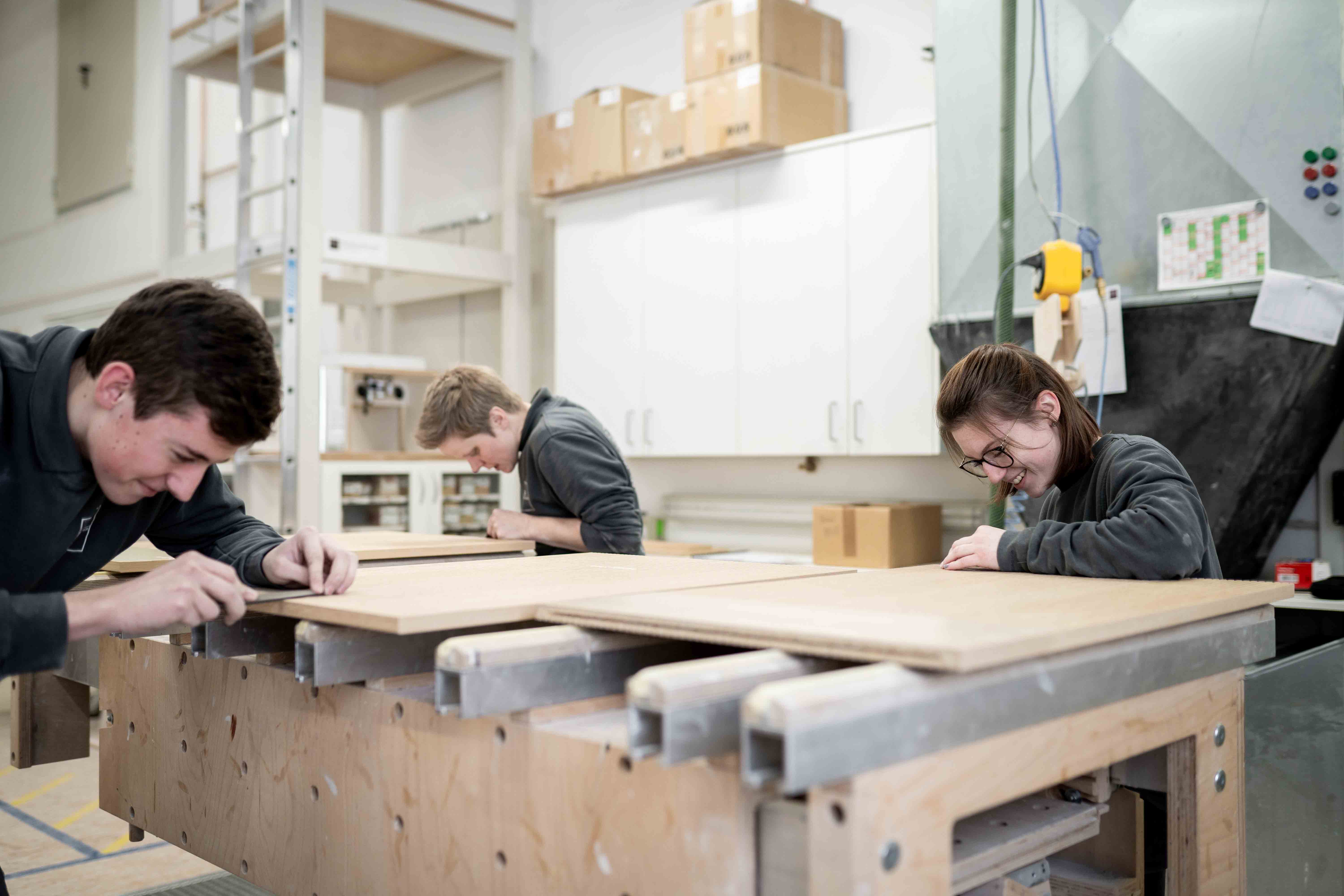 Drei Mitarbeitende bearbeiten Holzplatten gemeinsam an einer Werkbank