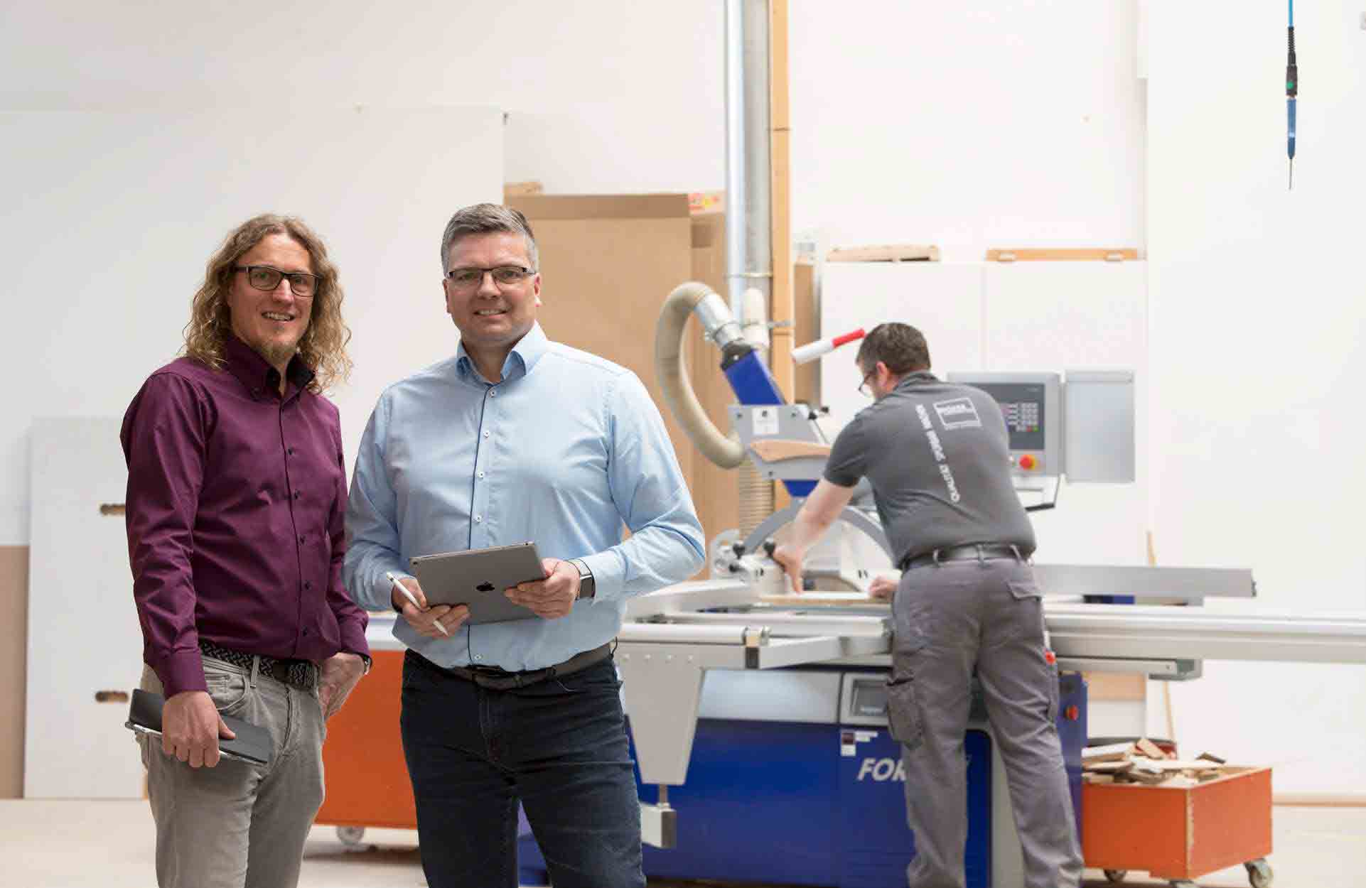 Zwei Geschäftsführer stehen mit Tablet in der Werkstatt, im Hintergrund arbeitet ein Mitarbeiter an einer Maschine