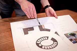 Person zeigt mit einem Stift auf einen ausgedruckten Raumplan auf einem Tisch