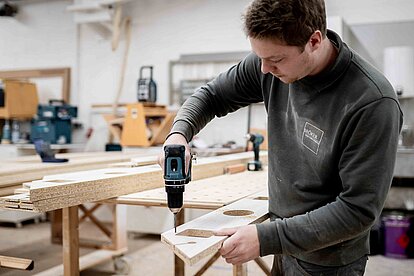 Mitarbeiter bohrt ein Loch in ein Holzbauteil an einer Werkbank